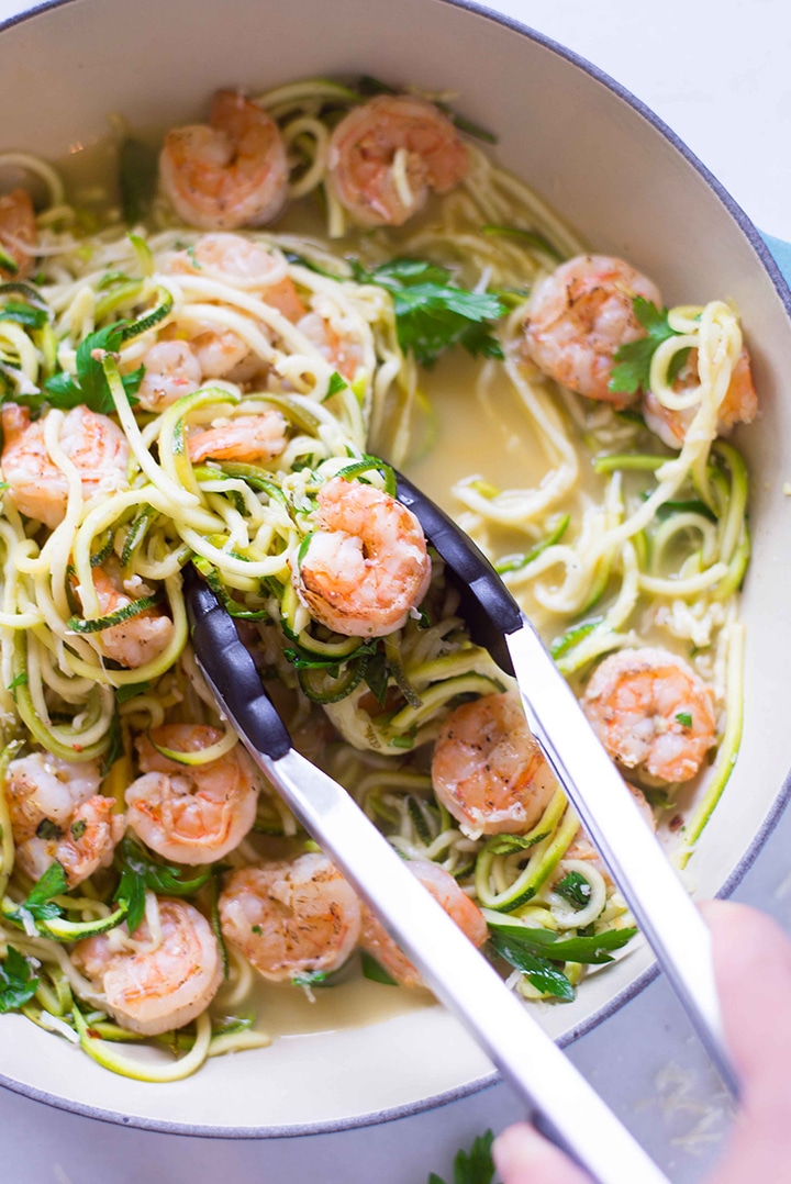 Large skillet with cooked zucchini noodles and shrimp scampi with a hand using a pair of tongs to grab some of the noodles and shrimp to serve.