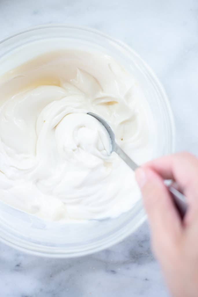 Image of a glass bowl containing the Greek yogurt frosting, with a hand holding a spoon dipping into the bowl.