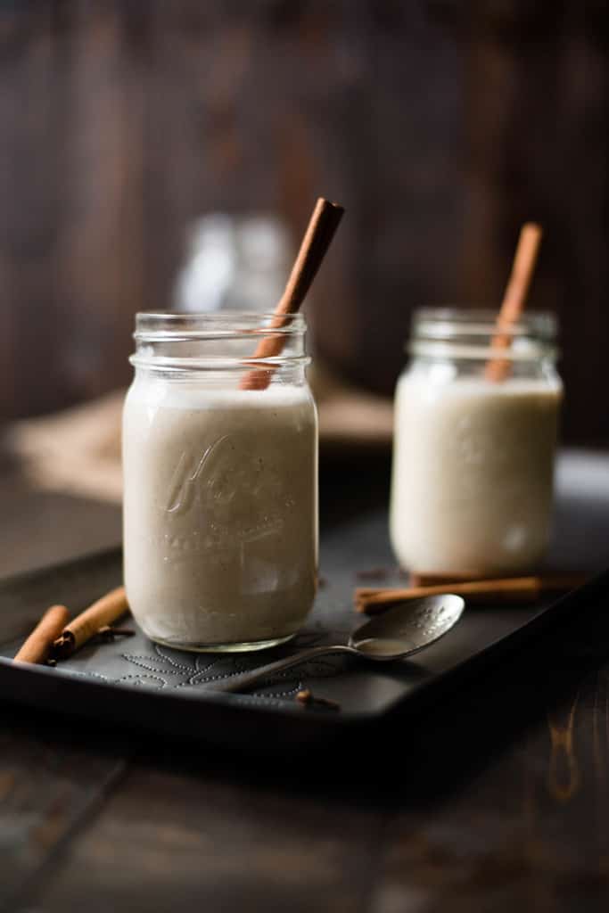 View of two mason jars filled with Non-Alcoholic Eggnog on a wooden tray, with cinnamon sticks in the eggnog.