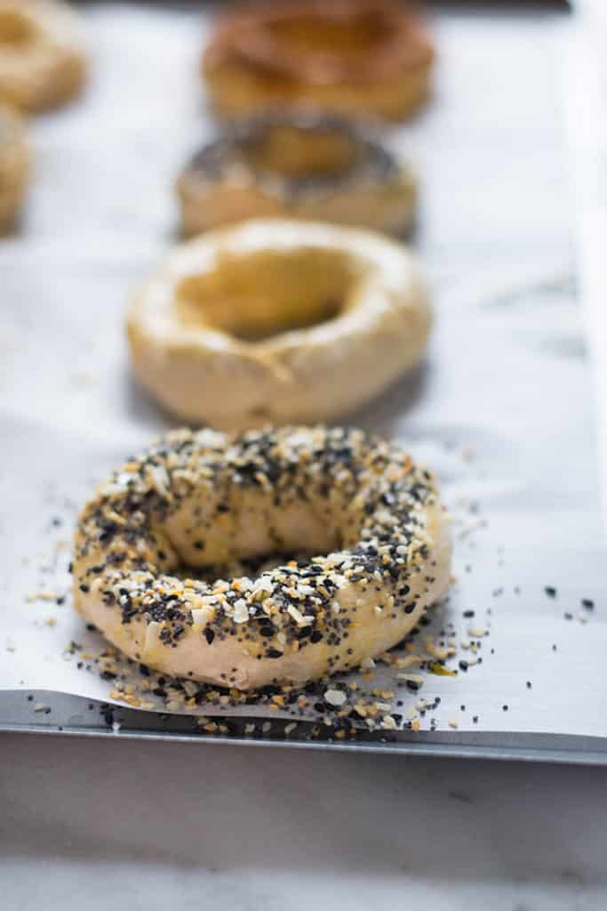 Side view of a baking sheet lined with parchment paper that has unbaked homemade bagels that are covered with various toppings, like everything mix and cinnamon sugar, that are ready to be baked for the homemade bagel recipe.