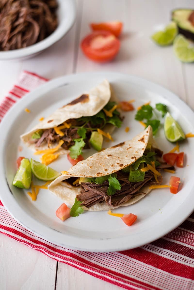 Overhead view of two Crock Pot Shredded Beef Tacos on a plate, including cheesem shredded tri-tip, and lime wedges.