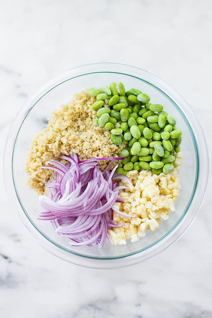 Overhead view of a mixing bowl, which contains shelled edamame, sliced red onions, cooked quinoa, and fresh corn, ready for the vinaigrette to be tossed in for the summer corn edamame salad