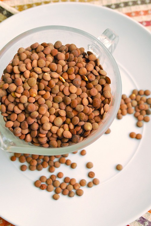 Overhead image of a clear glass bowl filled with dried lentils, resting on a white plate with a few lentils sprinkled around.