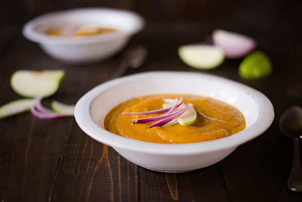 Horizontal image of two bowls of roasted acorn squash soup, with one bowl up close. Red onion and green apple slices are used as a garnish.