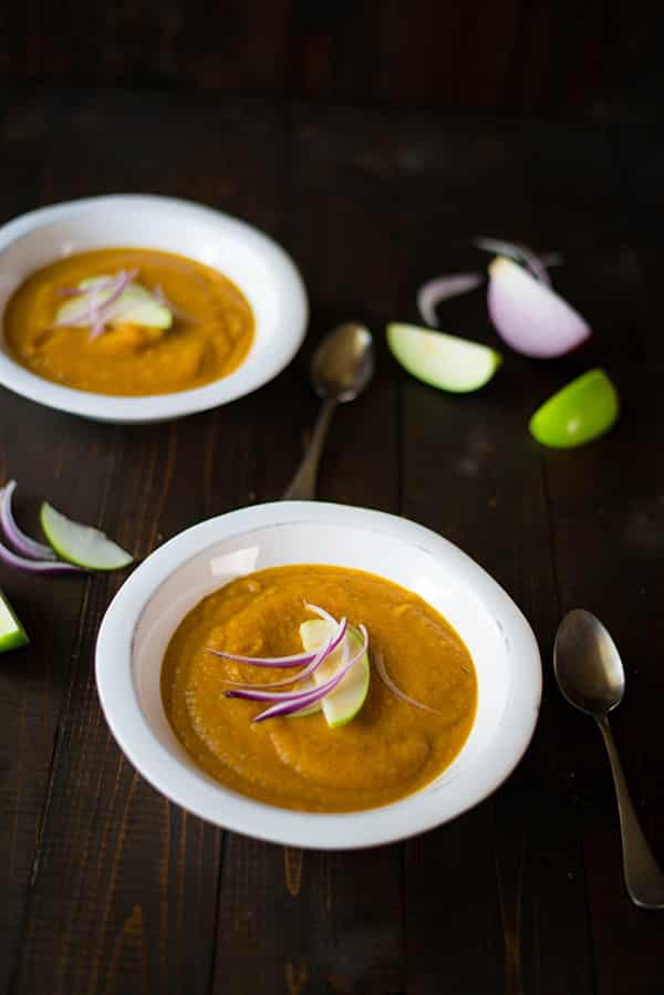 Overhead image of two bowls of roasted acorn squash soup. Next to the bowls are red onion and green apple wedges, which are also on top as a garnish.