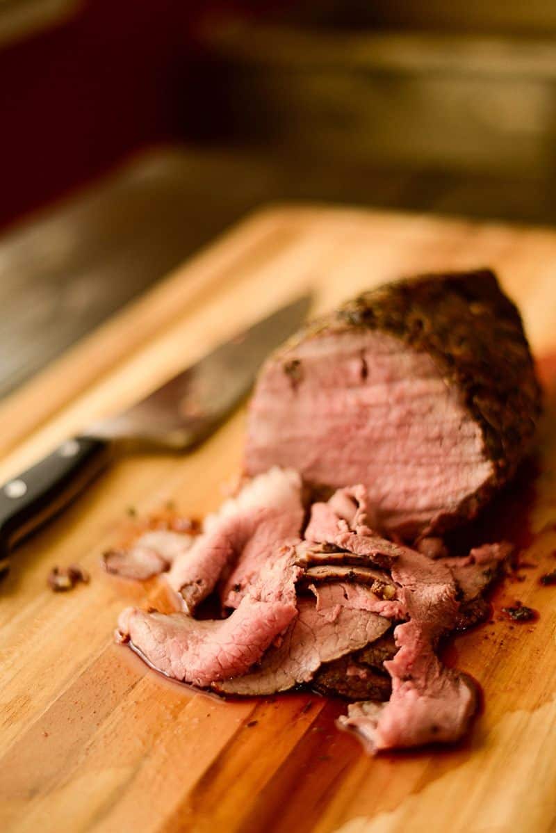 Close up image of Roast Beef partially sliced on a wooden cutting board with a knife laying beside it.