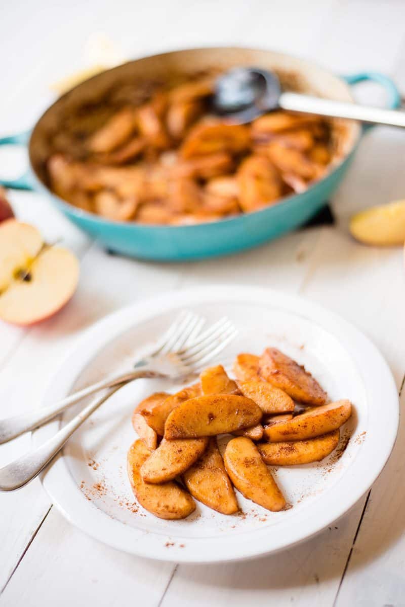 Side image of a blue skillet filled with cinnamon apples. In front of the skillet is a white plate full of cinnamon apples, with two forks beside it. 