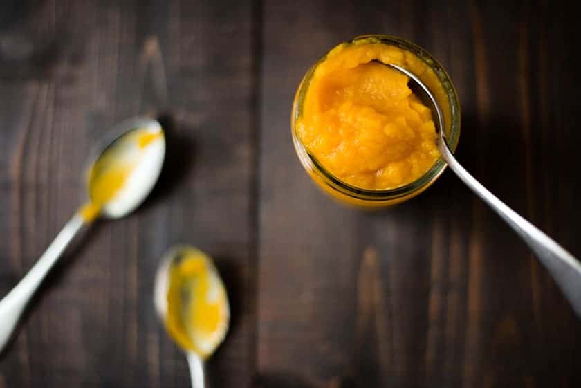 Overhead image of a glass jar filled with Pumpkin Puree From A Fresh Pumpkin, with a spoon dipping in it and 2 spoons beside the puree.
