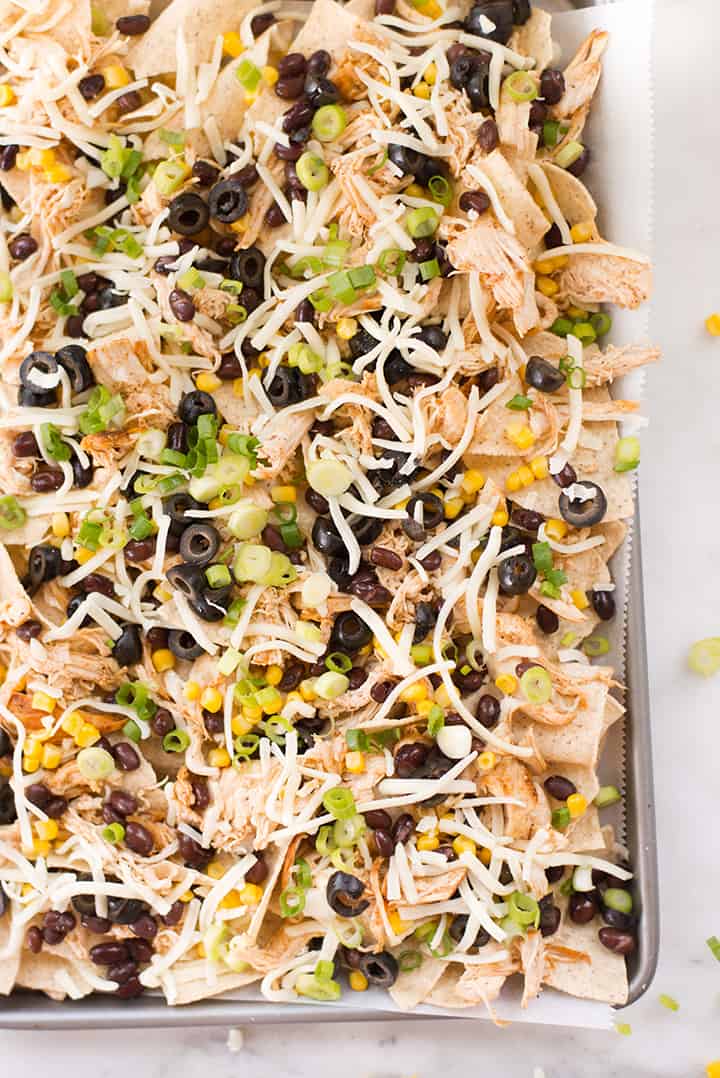 Overhead view of shredded chicken nachos on sheet pan with olives, black beans, green onions, shredded jack cheese, and other toppings, all ready to be baked.