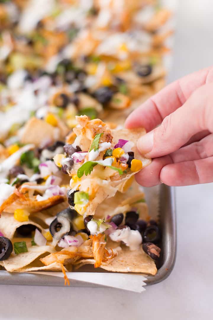Close-up of a hand holding a tortilla chip loaded with nachos toppings, ready to eat.