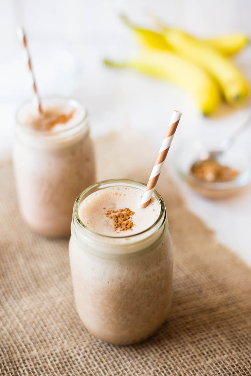 Side view of two mason jars filled with chocolate banana peanut butter protein shake, with bananas and peanut butter in the background.