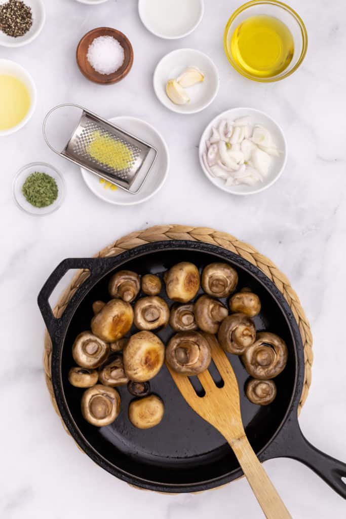 Overhead view of ingredients to make marinated mushrooms, including lemon, oregano, kosher salt, and garlic cloves, with a skillet of mushrooms nearby.