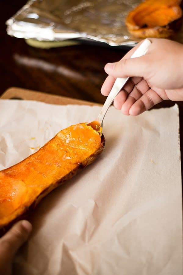 Hand holding a spoon instructing how to remove the roasted butternut squash from the skin after it has cooled from roasting in the oven.
