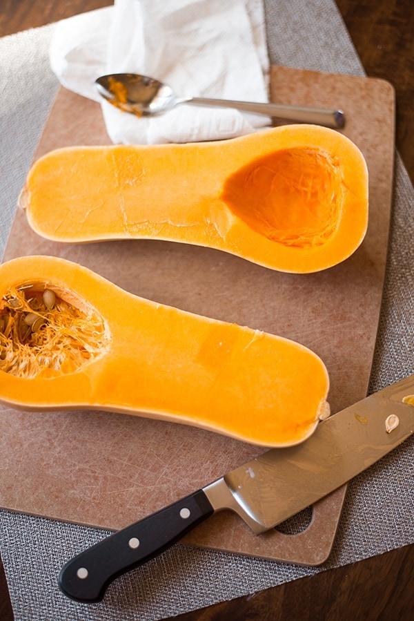 Cutting board with fresh butternut squash sliced in half with one side having all the seeds removed and other still needing the seeds and membranes to be removed in order to roast for the butternut squash bisque.