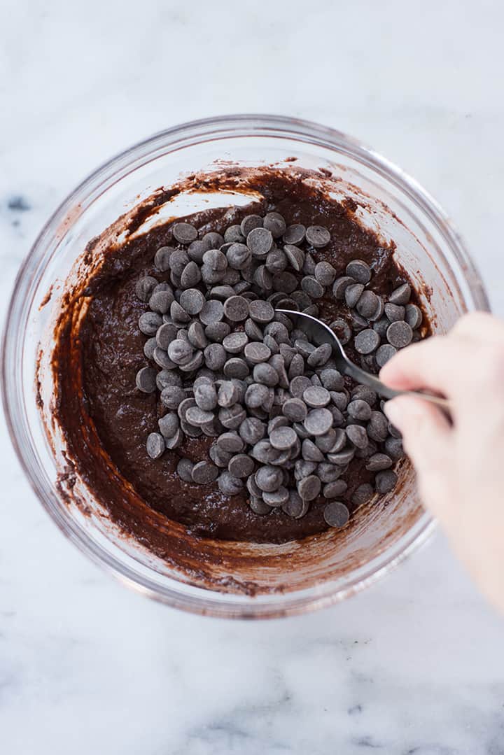 Overhead view of a glass bowl containing ingredients for Healthy Dark Chocolate Brownies, with the chocolate chips being folded in.