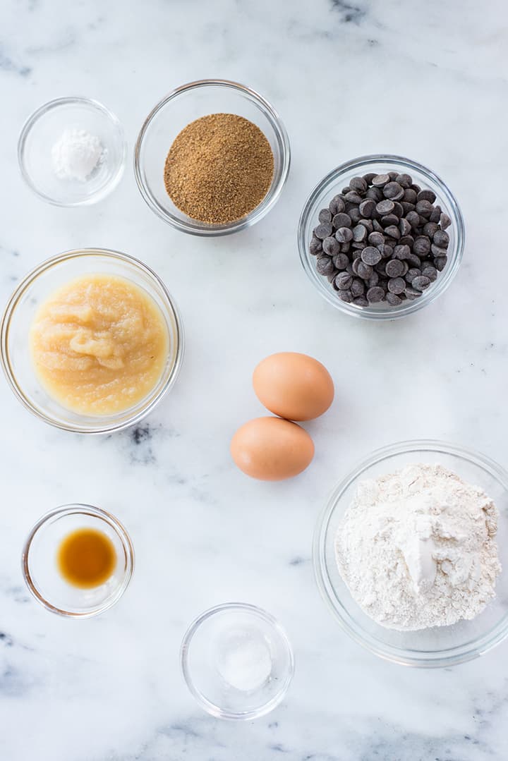 Overhead view of ingredients for Healthy Dark Chocolate Brownies, including whole wheat flour, dark chocolate chips, eggs, applesauce, baking powder, vanilla, coconut sugar and sea salt.