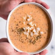 Two hands holding a white bowl of lobster bisque garnished with herbs and lobster meat, over a marble surface.