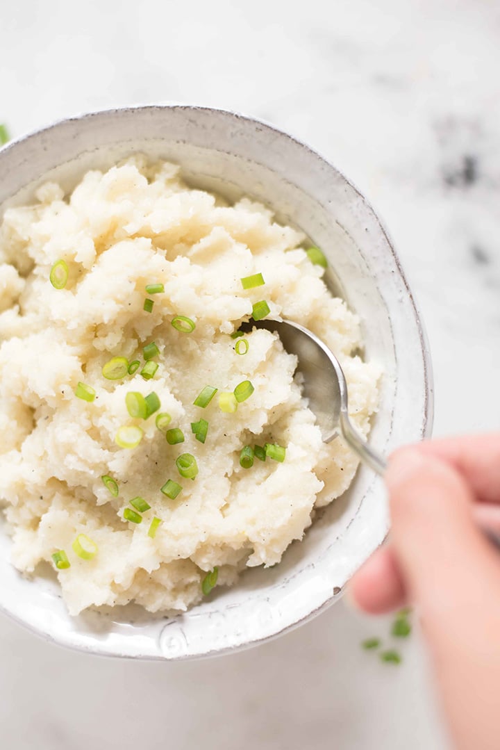 Cauliflower mashed potatoes in a bowl that are topped with sliced chives and ready to eat with a hand coming in with a spoon, about to eat.