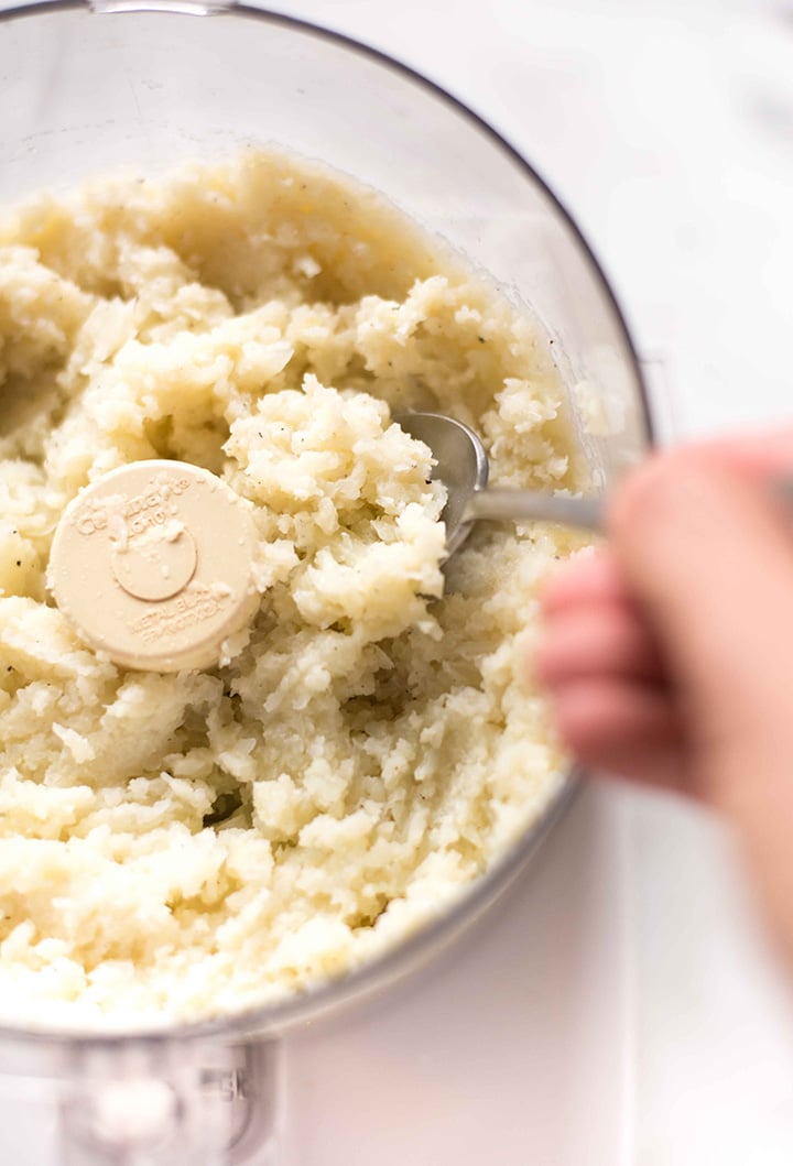 Close-up view of the pureed cauliflower mashed potatoes in the food processor with a spoon digging into the mixture to show the smooth texture.