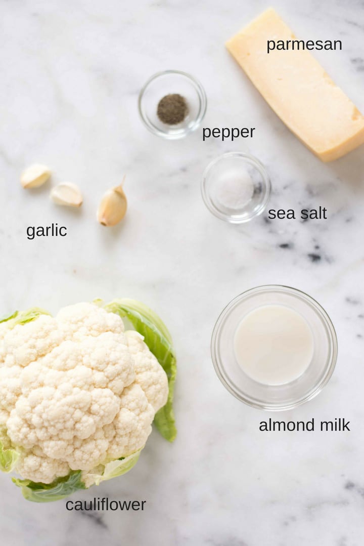 View of all the ingredients needed to make cauliflower mashed potatoes, including cauliflower, garlic, sea salt, pepper, parmesan cheese, and almond milk.
