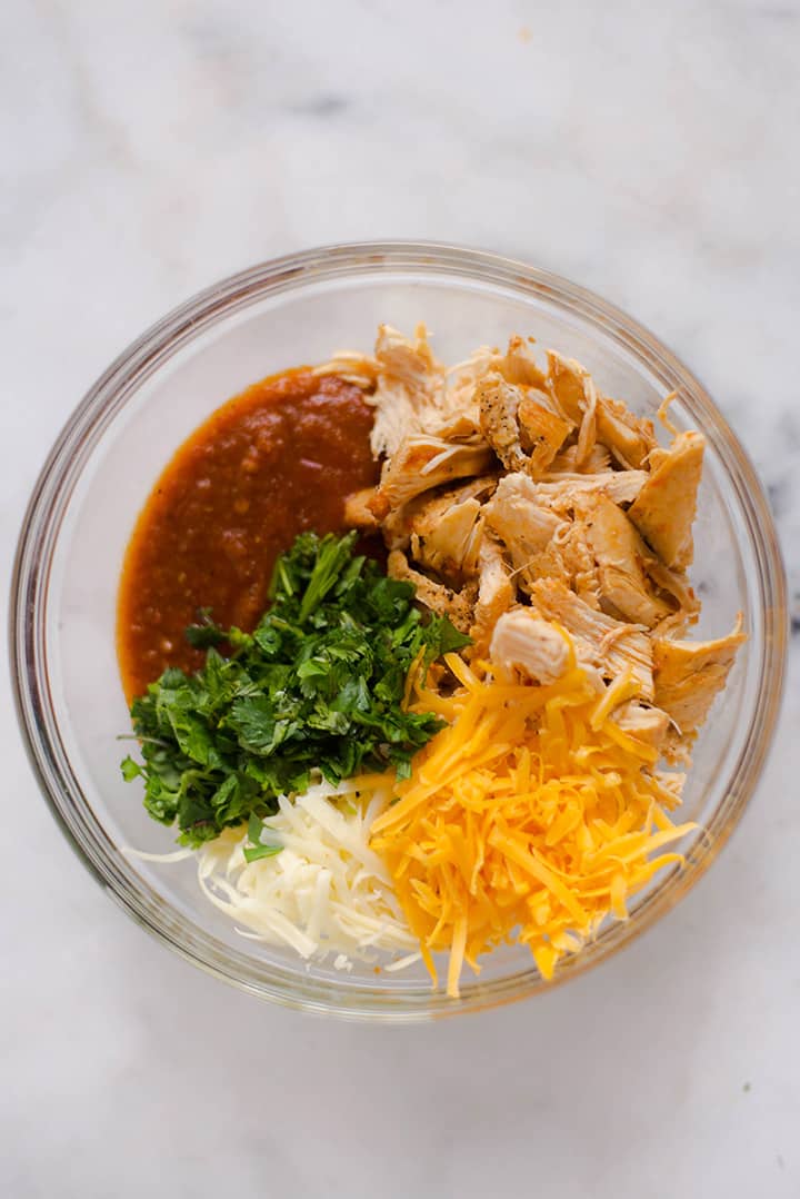 Overhead view of a mixing bowl which contains all the ingredients needed to make the filling for the shredded chicken enchiladas, including shredded chicken, homemade enchilada sauce, cilantro, jack and cheddar cheese.