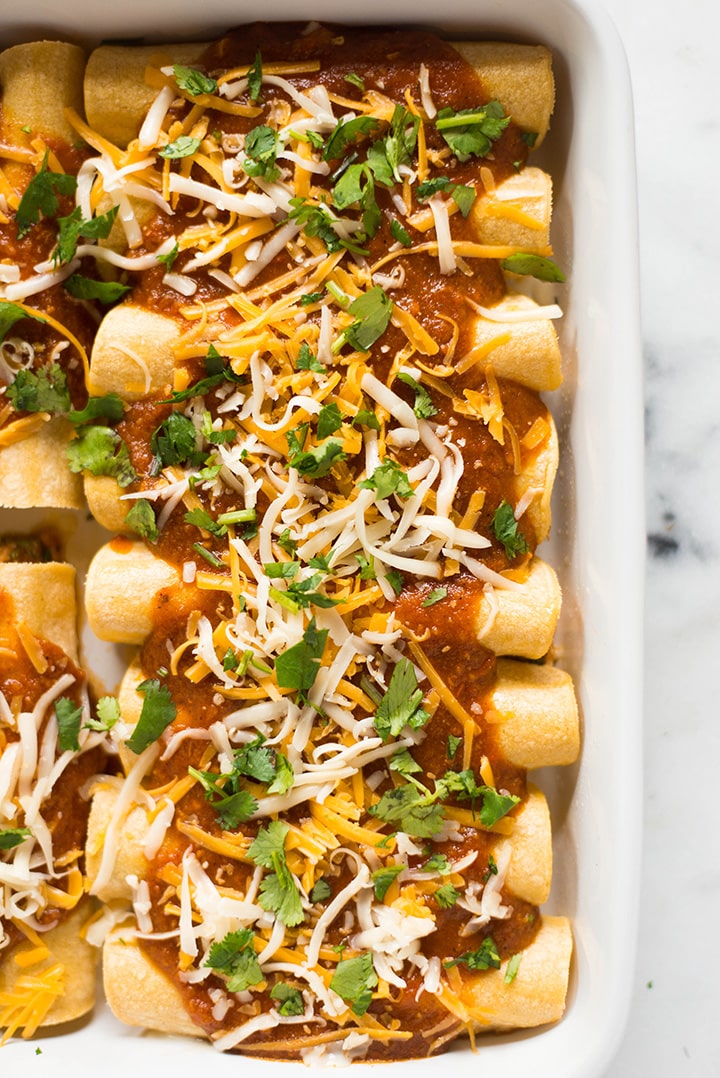 Overhead view of a casserole dish which is filled with shredded chicken enchiladas that have been topped with jack cheese, cheddar cheese, cilantro and red enchilada sauce, ready to bake.