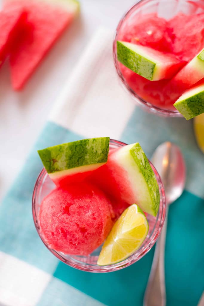 Overhead image of two glass cups filled with watermelon sorbet, along with fresh watermelon slices and lime wedges, ready to eat.