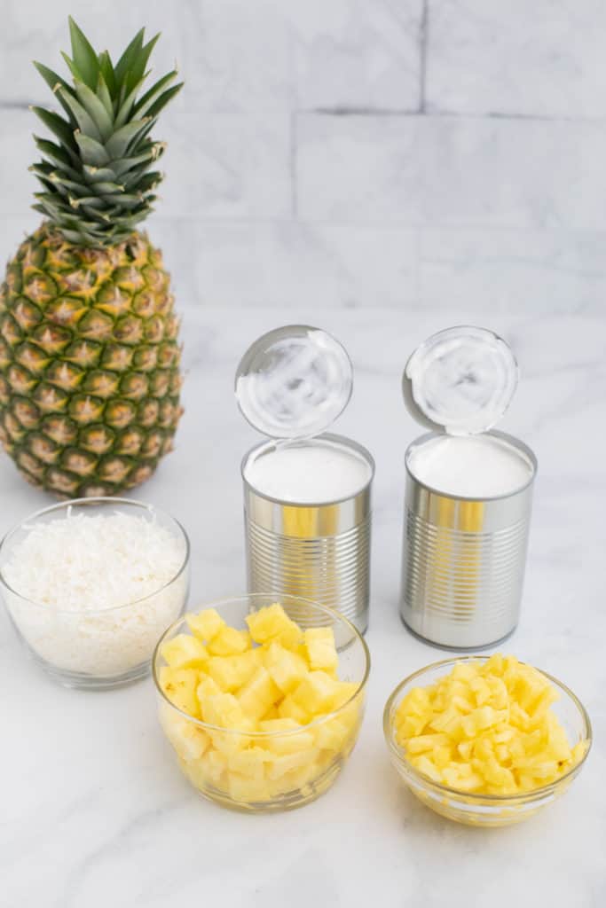Side view of bowls of pineapple, coconut, open cans of coconut milk and a whole pineapple nearby.
