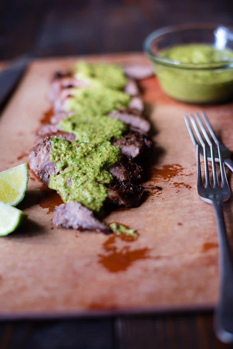 Close up side view of Flank Steak with Chimichurri Sauce, with a bowl of the sauce and two forks beside it.