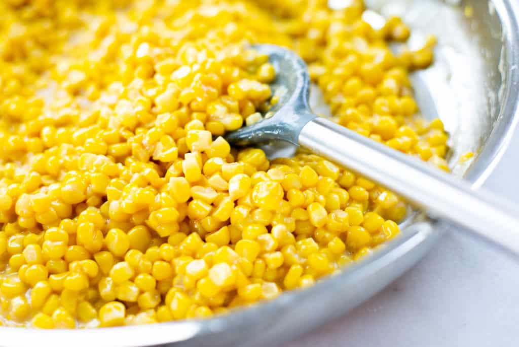 Close up view of a saucepan of fresh creamed corn, with a spoon resting in the saucepan.