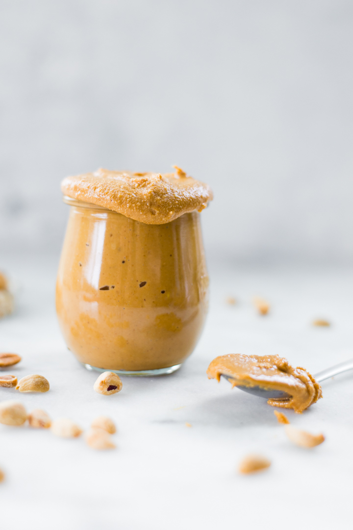 Side image of a glass jar full of homemade peanut butter, with a spoon of peanut butter beside it, as a source of vegan protein.