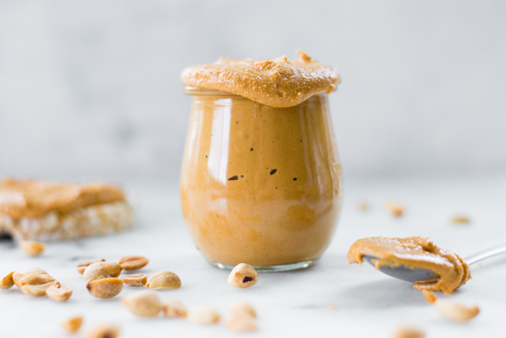 Close up image of a glass jar filled to the top with homemade honey roasted peanut butter, with peanuts sprinkled around the jar and a spoonful of peanut butter off to the side.