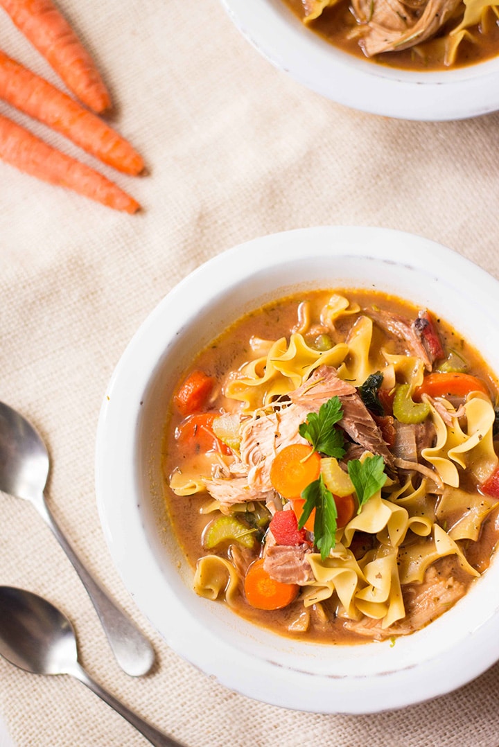 Overhead image of one bowl of easy turkey noodle soup that is topped with fresh Italian parsley and sitting next to raw carrots, which are also sliced and cooked into the soup.