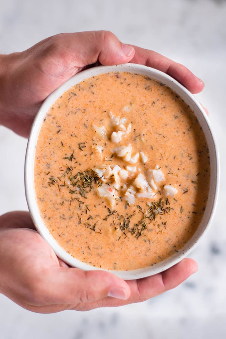 Hands holding one bowl filled with easy lobster bisque and topped with cooked lobster chunks and thyme.