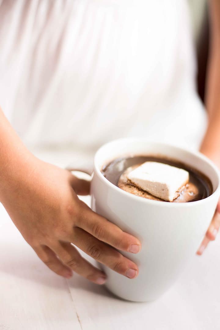 Girls hands cupping a white mug that has been filled with hot chocolate and topped with a homemade marshmallow.