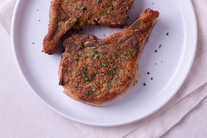Overhead image of two cooked Best Juicy Pork Chops being served on a white plate, which is on a white linen napkin.
