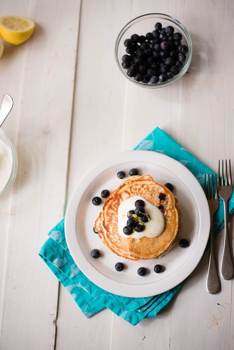 Overhead image of blueberry lemon high protein pancakes pictured with a bowl of fresh blueberries and lemons. Topped with greek yogurt topping. 
