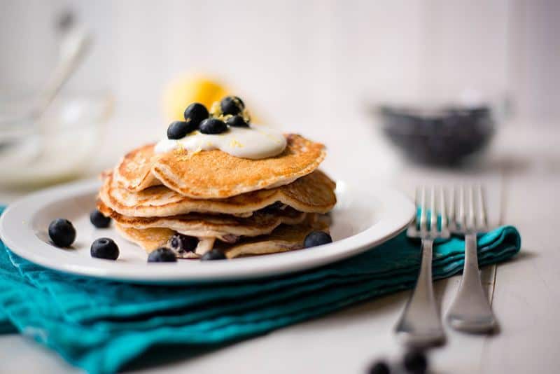 Side view of a stack of Lemon Blueberry High Protein Pancakes, topped with blueberries and on a white plate, ready to be enjoyed as a Healthy Breakfast Idea.