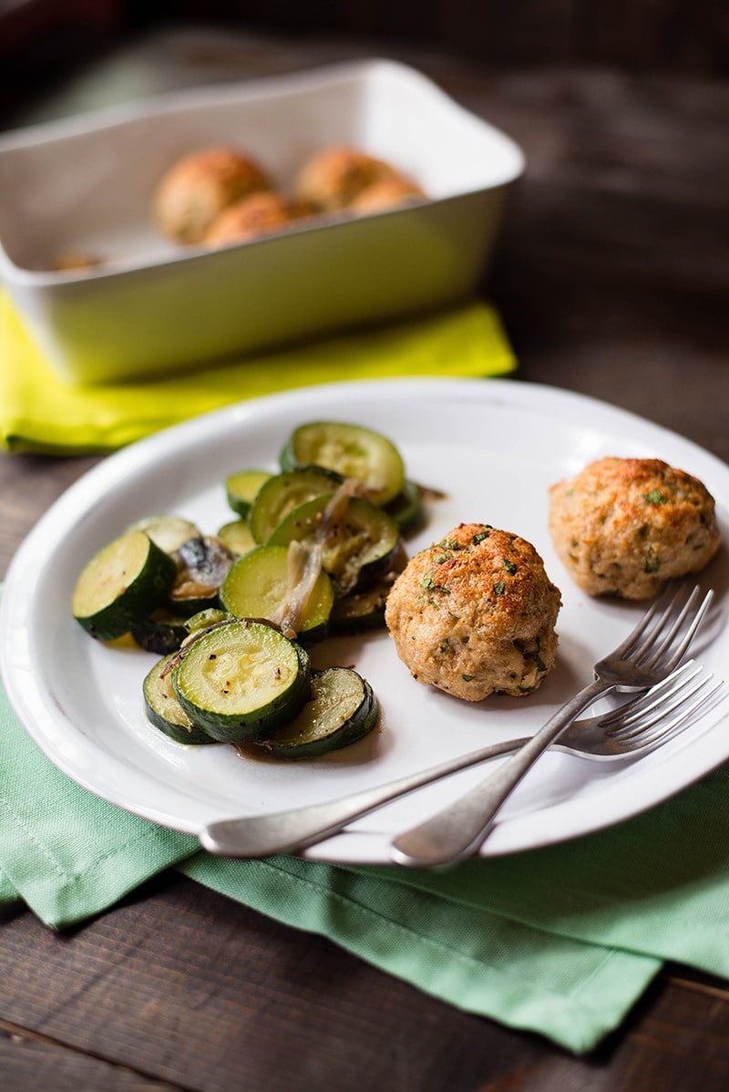 Healthy chicken meatballs on a plate next to sauteed zucchini. In the background are the baked chicken meatballs in a casserole dish.