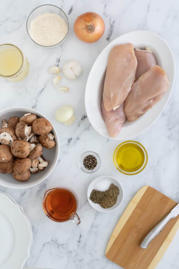 Overhead view of ingredients for Healthy Chicken Marsala laid out on the counter including mushrooms, garlic, chicken breasts, and onions.