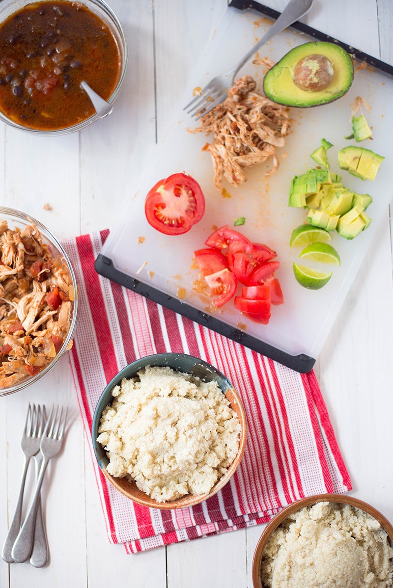 Bowls of shredded chicken, black beans, cauliflower rice, photographed with half an avocado, and diced tomatoes placed on a chopping board, all ready to assemble the slow cooker chicken burrito bowl with cauliflower rice