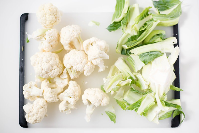 A cutting board with fresh cauliflower florets separated from the leaves of the cauliflower for how to make cauliflower cheese sauce.