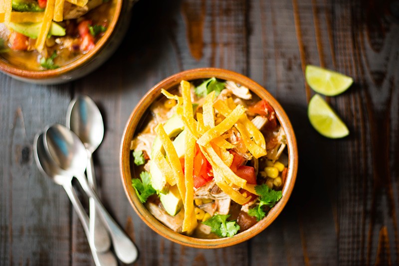 One bowl of slow cooker chicken tortilla soup, topped with tortilla strips, avocado, and fresh tomato, and next to fresh lime wedges for serving.