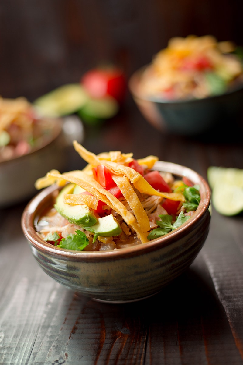 Close up image from the side angle of a bowl of slow cooker chicken tortilla soup. Can see the sliced avocado, tortilla strips, cilantro and chicken up close.