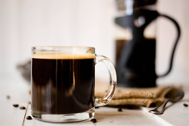 Close up side view of a glass mug containing black coffee, with a French press in the background.