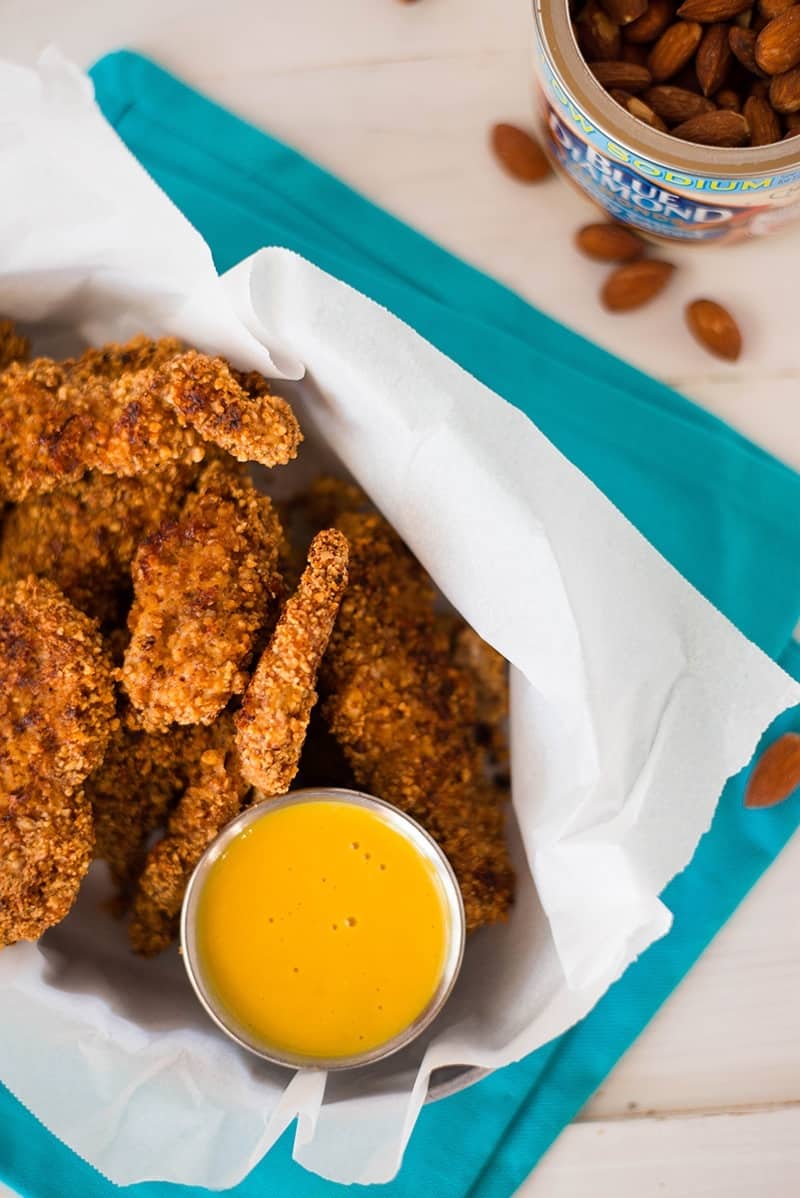 Close up photo of baked almond chicken strips placed on oil-absorbent cooking paper, together with honey mustard dipping sauce and a can of lightly salted almonds 