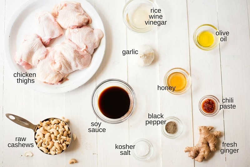 All the ingredients for the slow cooker cashew chicken, separated and ready to add to the slow cooker. Each ingredient has a label, including chicken thighs, low sodium soy sauce, raw cashews, rice wine vinegar, honey, garlic, salt, pepper, fresh ginger, olive oil, and chili paste.