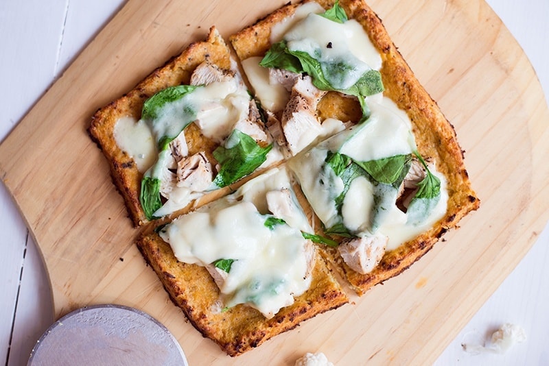Cauliflower pizza crust topped with seasoned chicken breast, fresh spinach, and mozzarella cheese, freshly baked, and setting on a wooden pizza paddle.