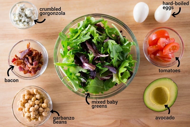 All the ingredients placed in a bowl, which are used to make a steak Cobb salad, including crumbled gorgonzola, hard boiled eggs, bacon, garbanzo beans, mixed baby greens, tomatoes, and avocado