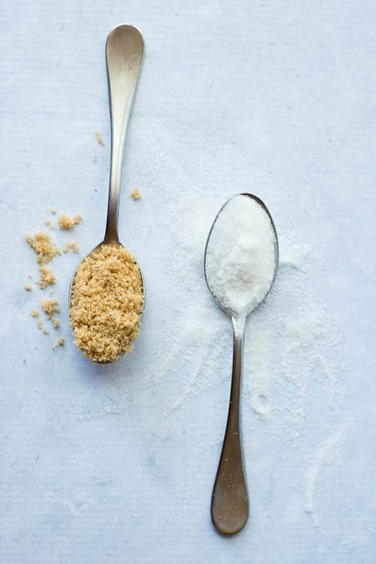 Overhead image of two teaspoons filled with sugar, one white sugar and the other brown sugar.
